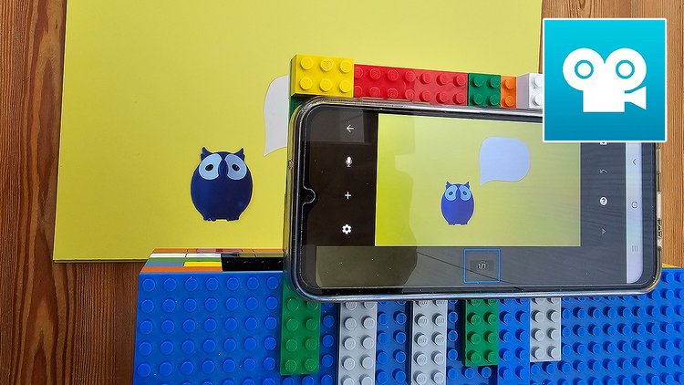Auf dem Bild ist ein Smartphone auf einem selbst gebastelten Lego-Halter zu sehen, das eine Eule auf Papier fotografiert. Dazu sieht man rechts oben das Logo der App Stop Motion Studio