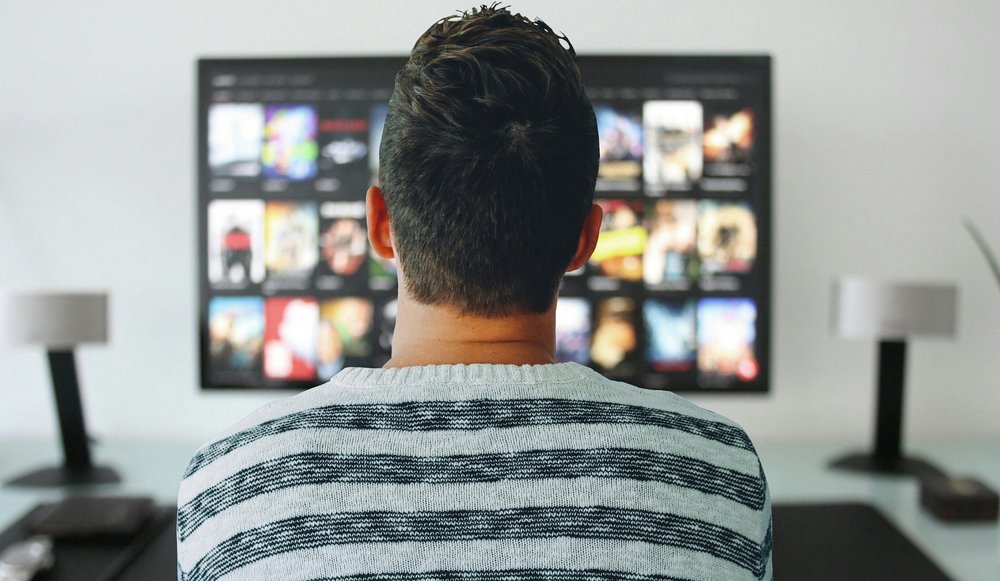 Ein Mann sitzt vor einem Fernseher, am Fernseher sind vage verschiedene Filmcover-Bilder zu erkennen.