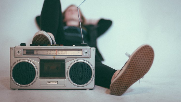 Im Vordergrund steht ein Radio, dahinter liegt entspannt ein junger Mensch und hört zu.