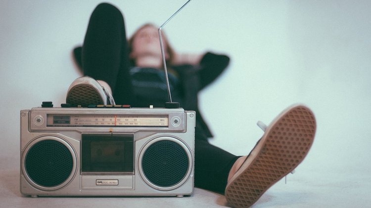 Im Vordergrund steht ein Radio, dahinter liegt entspannt ein junger Mensch und hört zu.