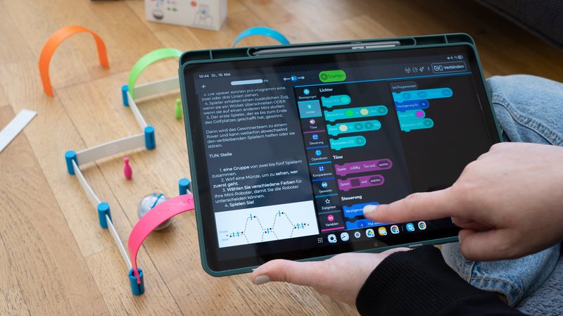 Nahaufnahme eines Tablets, auf dem die Programmieroberfläche der Sphero Edu App geöffnet ist. Im Hintergrund am Boden ist ein Sphero Mini-Parcours und der Sphero Mini-Roboter zu sehen.