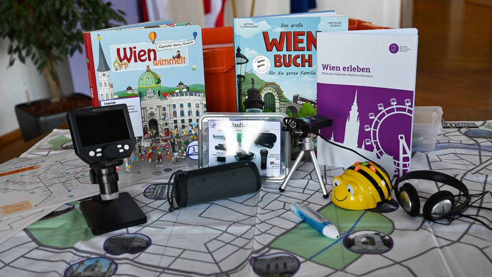 Inhalte der verschiedenen Digiboxen sind auf einem Tisch aufgestellt: Wien-Matte, Bee-Bot, Audioaufnahmegerät, Bücher, ein digitales Mikroskop, ein "Wien erleben"-Booklet und einiges mehr.