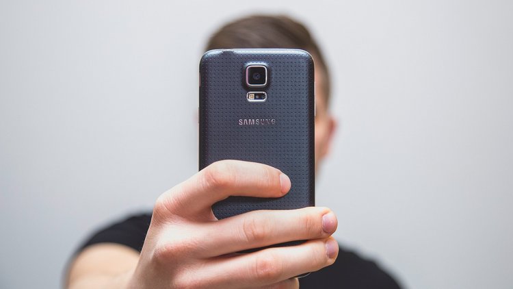 Ein junger Mann hält ein Smartphone in der Hand. Das Gesicht des Mannes ist durch das Smartphone verdeckt, es wirkt als würde er ein Foto von sich selbst machen.