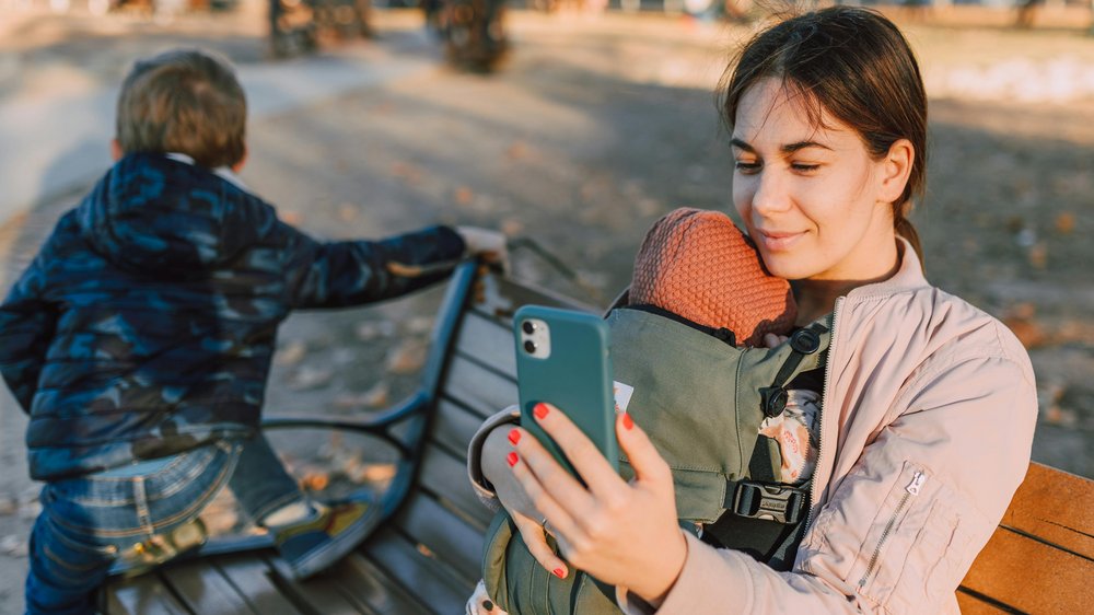 Eine Frau mit Baby in einem Tragegurt sitzt bei herbstlicher Stimmung auf einer Bank und hält ein Smartphone vor sich.