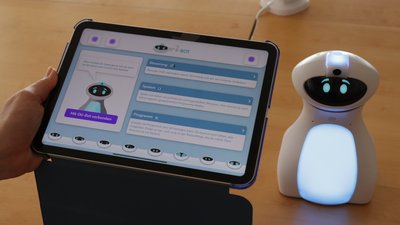 Eine Hand hält ein Tablet, darauf ist die Oberfläche der Oti-Bot-App zu sehen. Im Hintergrund steht der eingeschaltene Oti-Bot auf einem Tisch.