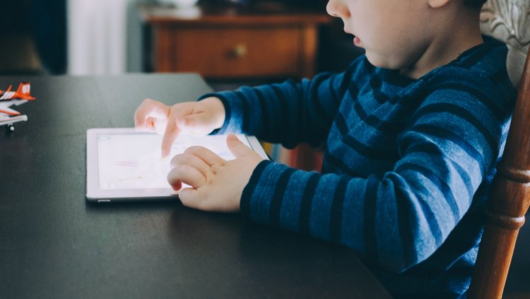 Auf einem Tisch liegt ein Tablet. Ein junges Kind sitzt davor und schaut rein.