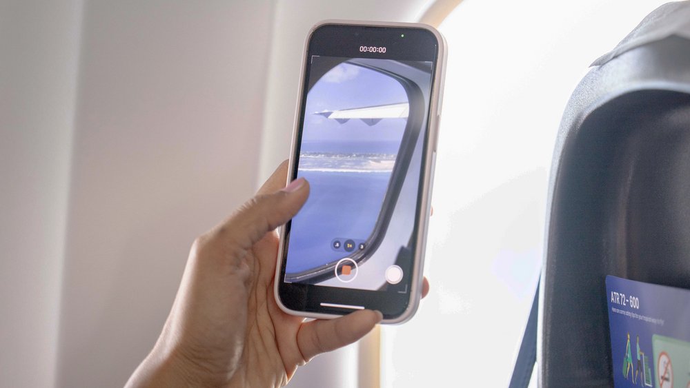 Auf dem Bild ist eine Hand, die ein Smartphone hält, zu sehen. Die dahinterstehende Person macht offenbar gerade ein Bild aus einem Flugzeug durch die Seitenfenster auf die Flugzeugflügel und darunterliegende Inseln.