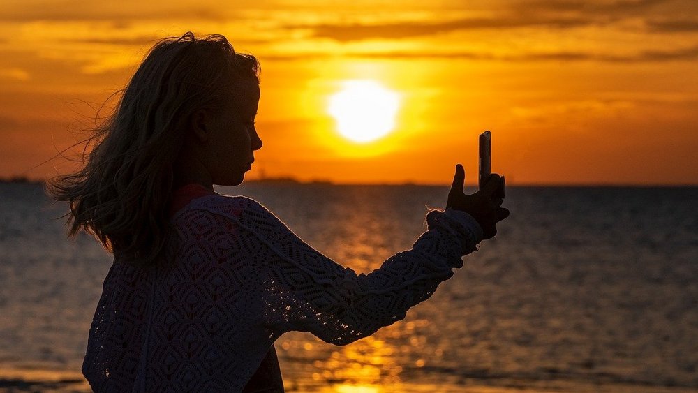 Im Hintergrund ist ein Sonnenuntergang am Meer zu sehen, davor ist die dunkle Silhouette eines Mädchens mit einem Smartphone in der Hand zu erkennen, das scheinbar gerade ein Selfie aufnimmt.