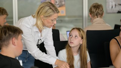 Schüler:innen sitzen an einem Tisch, eine erwachsene Frau beugt sich zu ihnen hinunter und erklärt etwas.