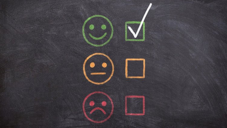 Auf einer Schultafel sind drei verschiedene Emojis neben 3 Checkboxen aufgezeichnet, ein freundlich gestimmter, ein mittelmäßig gestimmter und ein traurig gestimmter Emoji. Die Checkbox neben dem freundlichen Emoji ist abgehakt.